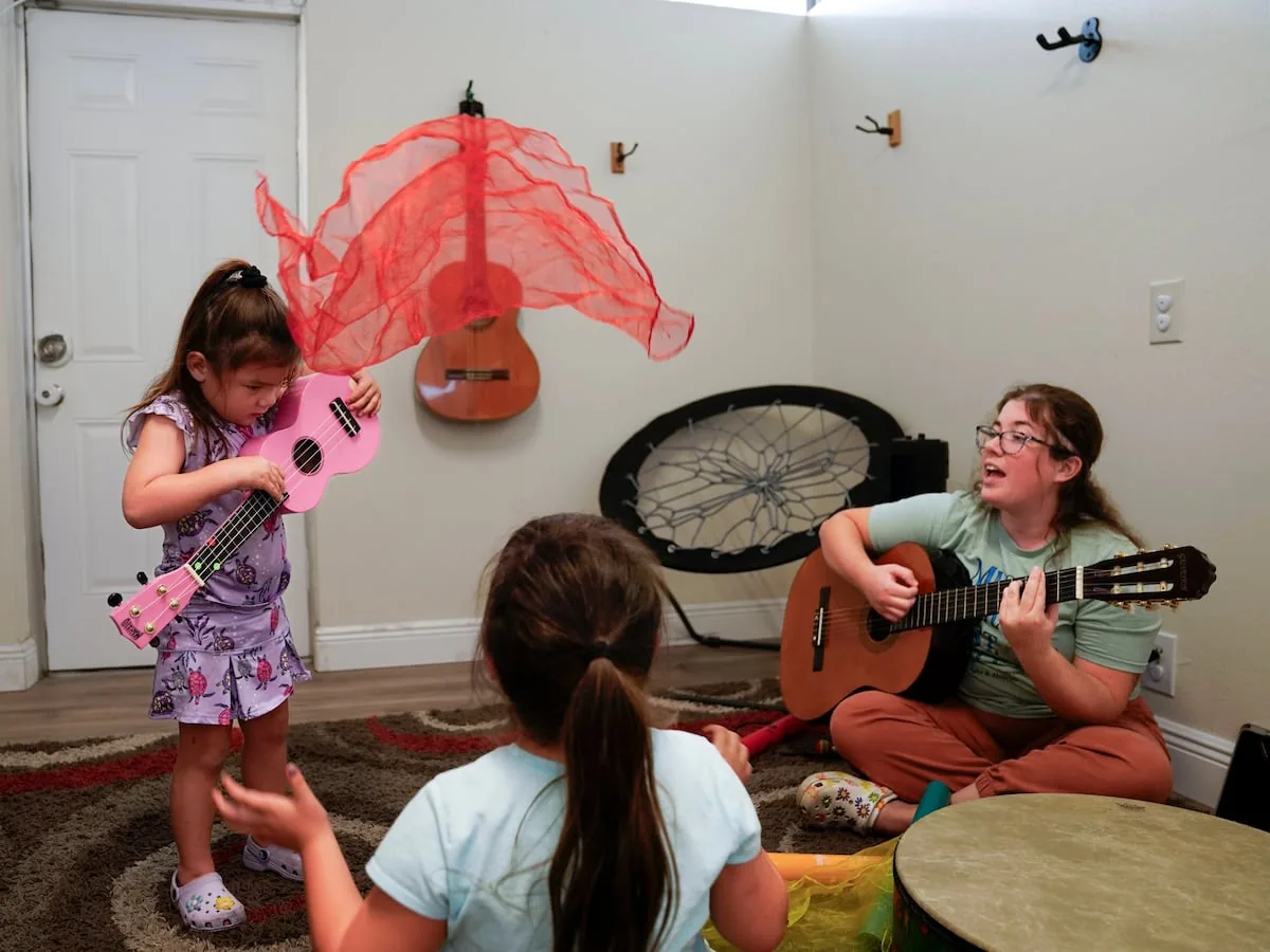 A board-certified music therapist plays guitar one-to-one with a child holding a ukulele and a movement scarf.