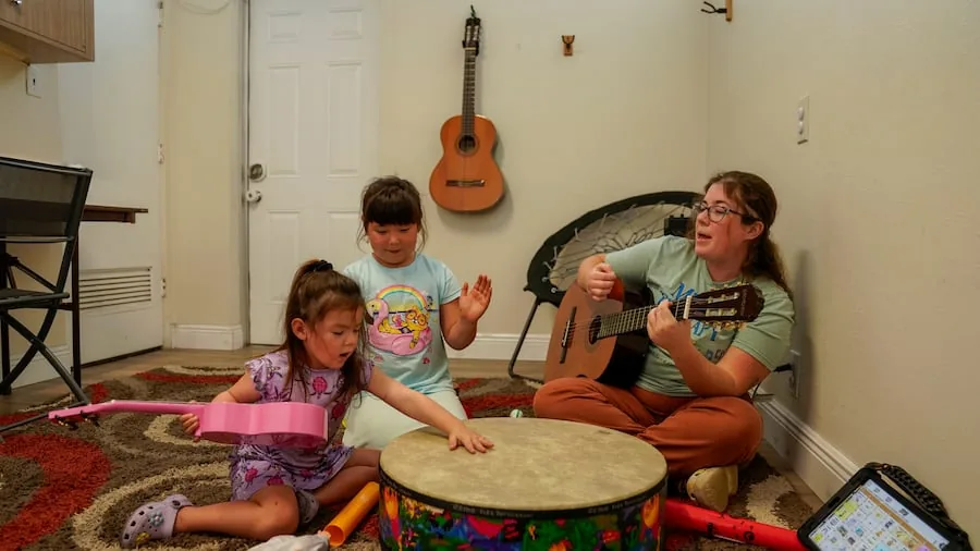 A board-certified music therapist plays guitar while leading an in-home session with two young children and a drum and ukulele.