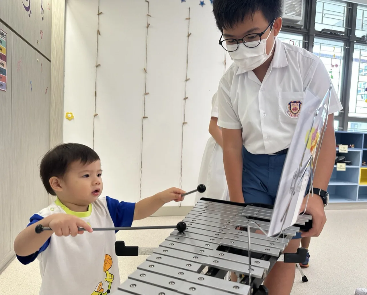 An older child helps a toddler tap a xylophone together.